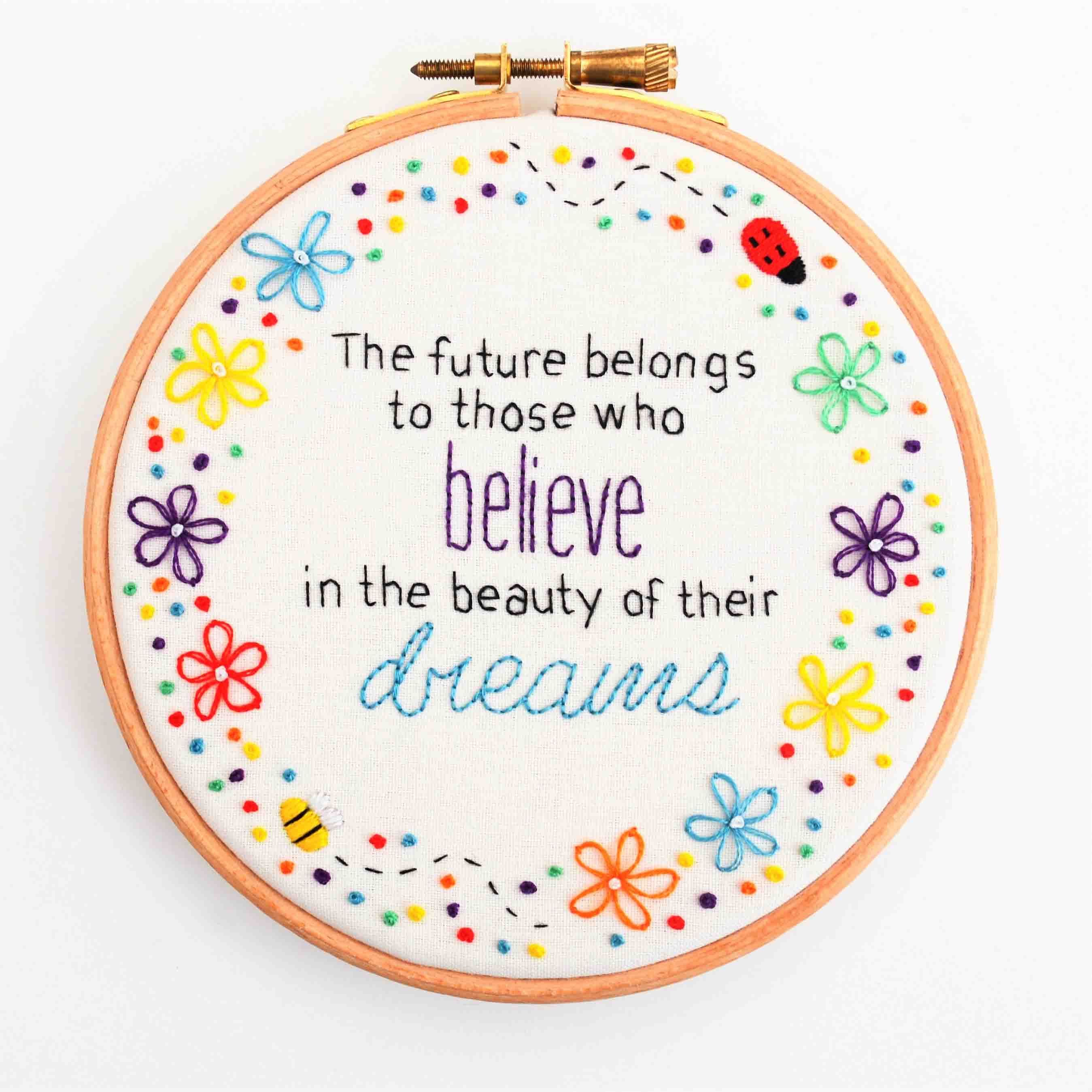 A white hoop with a wooden frame embroidered with a multicoloured polka dot border interspersed with multicoloured flowers, a ladybird and a bee with the words 'The future belongs to those who believe in the beauty of their dreams' in the middle