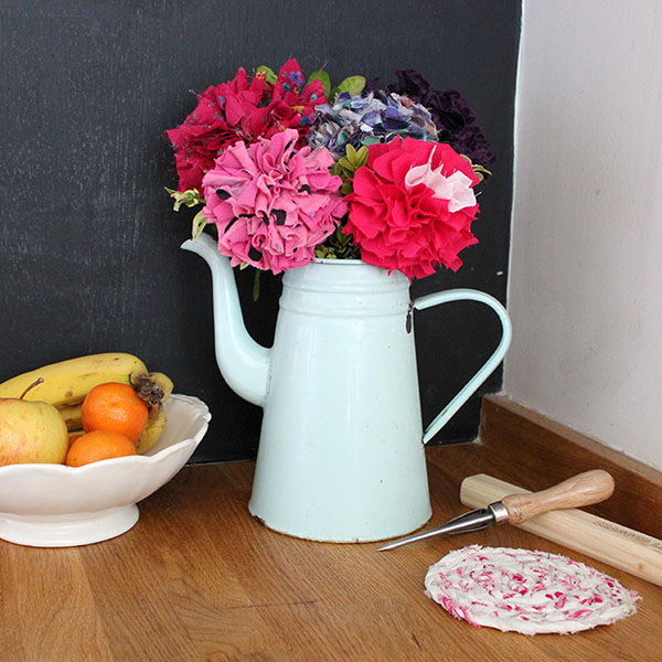 A bunch of pink, red and purple rag flowers in a white teapot on a table next to a fruit bowl