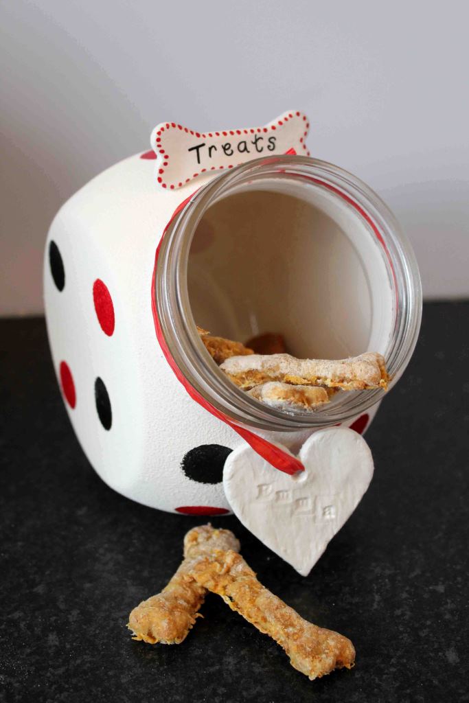 A white jar decorated with red and black spots placed on its side. At the top of the opening is a sign in the shape of a bone with the word 'treats' and a white heart shape is tied to the bottom with a ribbon. A few dog biscuits are spilling out of the jar.