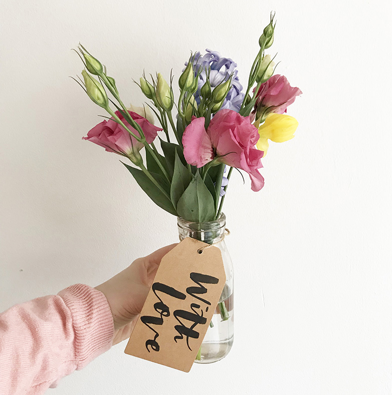 A posy of pink, purple and yellow flowers in a glass vase with a brown paper gift tag with the words 'With love