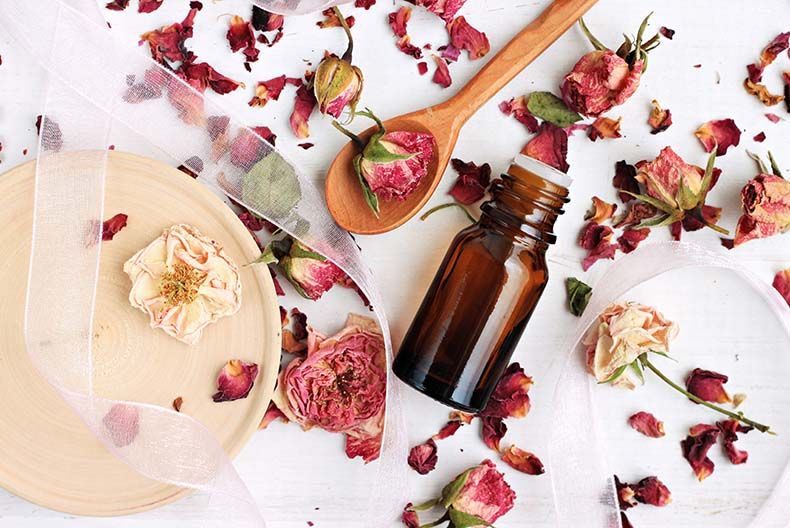 A pretty arrangement of dried rose petals and rose heads scattered across a white surface along with a wooden spoon and small toiletries bottle and some white lace draped over it