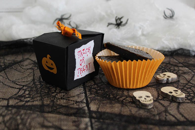 A black card box decorated with a smily pumpkin face on one side and the words 'Trick or treat' written in red with drops of blood falling off them on a white piece of paper on the other, tied with an orange ribbon, positioned next to an orange cupcake case filled with two treacle toffees on a piece of black lace with a spider web pattern