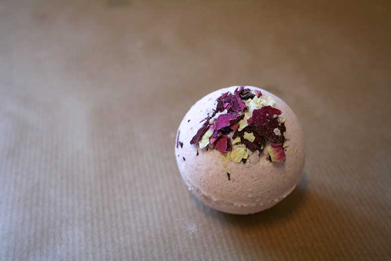 A light pink bath bomb adorned with purple and white petals on a brown paper surface