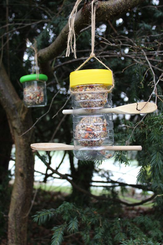 A plastic bird feeder with a yellow lid hanging from a tree with another plastic bird feeder with a green lid in the background